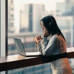Young woman with headphones using a laptop by a city window, engaged in remote work.