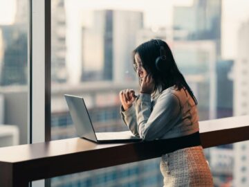 Young woman with headphones using a laptop by a city window, engaged in remote work.