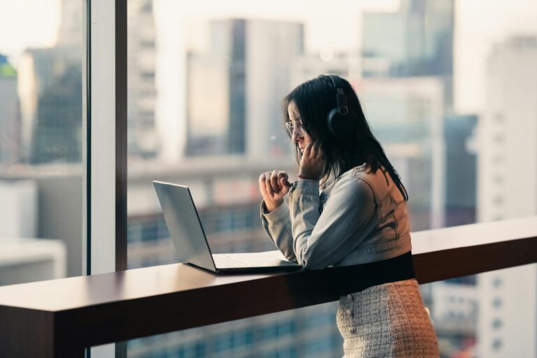 Young woman with headphones using a laptop by a city window, engaged in remote work.