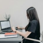 Woman working on laptop at desk with notebooks.