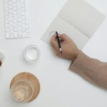 A top view of a minimalist workspace featuring a person writing, keyboard, and decor items.