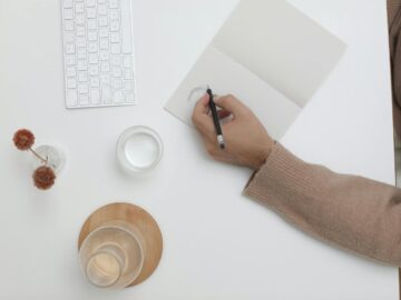 A top view of a minimalist workspace featuring a person writing, keyboard, and decor items.