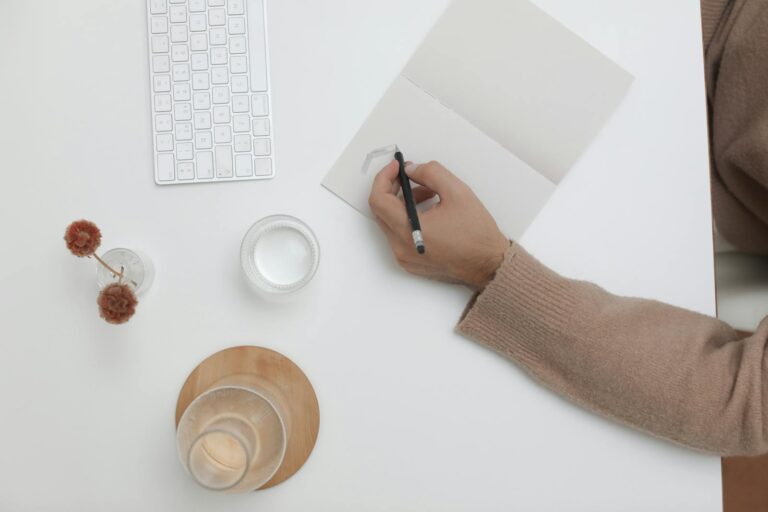 A top view of a minimalist workspace featuring a person writing, keyboard, and decor items.