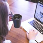 A woman enjoys coffee while browsing the web on a laptop at a home office desk.