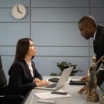 Two colleagues in a formal business setting engaged in a discussion at an office desk.
