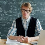 Man in glasses sits at a desk with chalkboard.