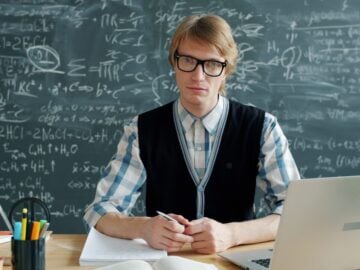 Man in glasses sits at a desk with chalkboard.