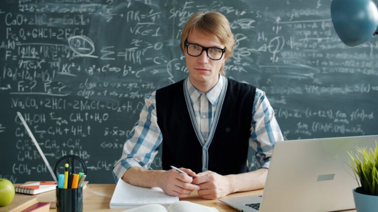 Man in glasses sits at a desk with chalkboard.