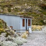 Small corrugated metal cabin in a rocky, overgrown landscape.