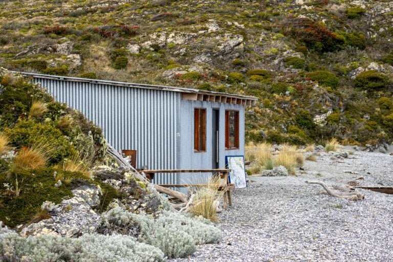 Small corrugated metal cabin in a rocky, overgrown landscape.