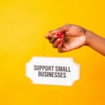 Hand holding a 'Support Small Businesses' sign against a vibrant yellow background, promoting local commerce.