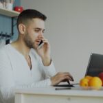 Man talking on phone while working on laptop at table.
