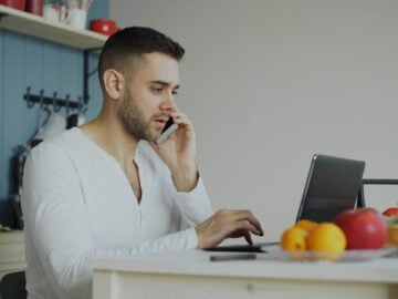 Man talking on phone while working on laptop at table.