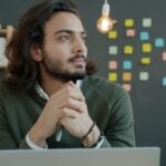 Man with long hair thinking at desk with laptop