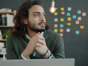 Man with long hair thinking at desk with laptop