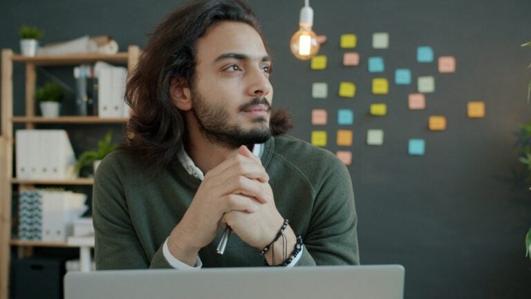 Man with long hair thinking at desk with laptop